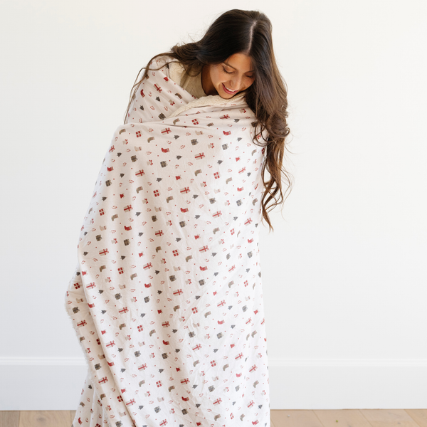 Woman wrapped in a holiday blanket with a festive pattern featuring gift boxes, stockings, Christmas trees, and other holiday illustrations. Ideal for cozy winter decor and holiday gifts that bring warmth and joy. The soft blanket is a luxury blanket and a Christmas blanket. 