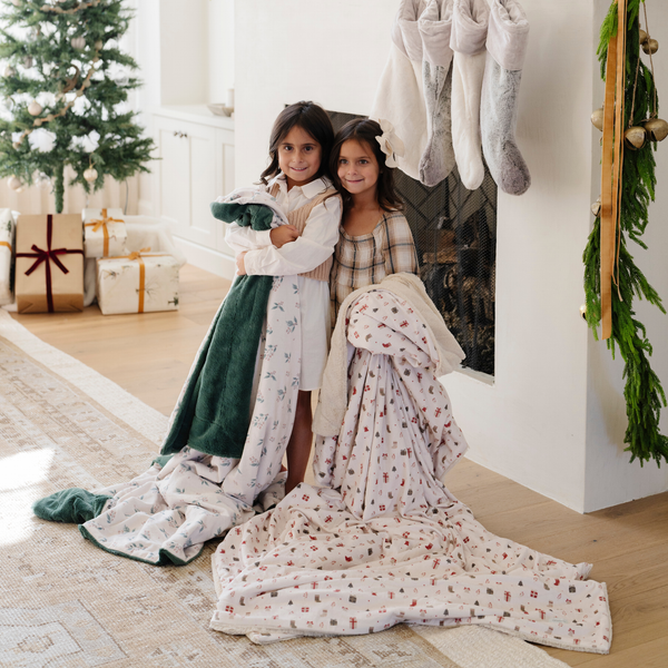 Two girls standing by a fireplace, wrapped in holiday-themed blankets with Christmas decor in the background. The scene includes a decorated tree, wrapped presents, and stockings, perfect for capturing the holiday spirit with cozy throw blankets. One blanket features a holly berry print, and the other has a holiday-themed pattern with gift boxes. Ideal for holiday gifting and cozy winter moments. The soft blankets are luxury blankets and Christmas blankets. 