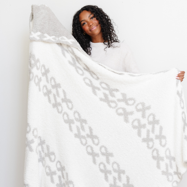 A smiling woman holds up a luxurious double-layer Bamboni® blanket by Saranoni, featuring a white and gray blanket with a cancer ribbons pattern on it. This ultra-soft blanket is crafted from bamboo rayon and poly microfiber, making it lightweight, breathable, and fluffy. Ideal as a cozy throw blanket, luxury blanket, plush throw, soft blanket, or stylish home accent, this Saranoni blanket brings warmth, comfort, and timeless elegance to any living room or bedroom.