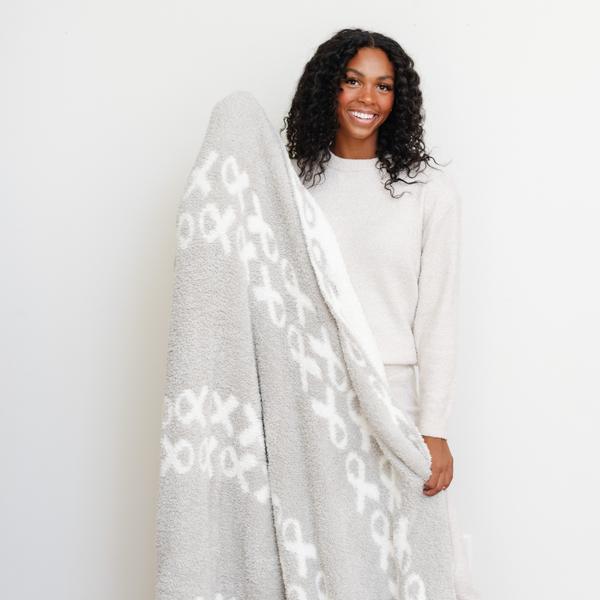 A smiling woman holds a luxurious double-layer Bamboni® blanket by Saranoni, featuring a white and gray blanket with a cancer ribbon pattern on it. This ultra-soft blanket is crafted from bamboo rayon and poly microfiber, making it lightweight, breathable, and fluffy. Ideal as a cozy blanket, extra large blanket, luxury blanket, soft blanket, or stylish home accent, this Saranoni blanket brings warmth, comfort, and timeless elegance to any living room or bedroom.