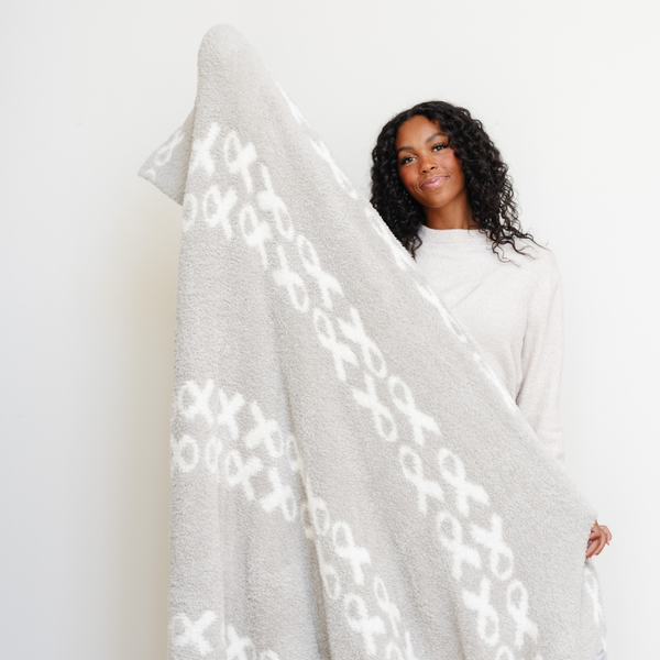 A smiling woman holds up a luxurious double-layer Bamboni® blanket by Saranoni, featuring a white and gray blanket with a cancer ribbon pattern on it. This ultra-soft blanket is crafted from bamboo rayon and poly microfiber, making it lightweight, breathable, and fluffy. Ideal as a cozy blanket, extra large blanket, luxury blanket, soft blanket, or stylish home accent, this Saranoni blanket brings warmth, comfort, and timeless elegance to any living room or bedroom.