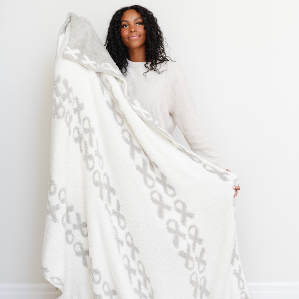 A woman holds a luxurious double-layer Bamboni® blanket by Saranoni, featuring a white and gray blanket with a cancer ribbon pattern on it. This ultra-soft blanket is crafted from bamboo rayon and poly microfiber, making it lightweight, breathable, and fluffy. Ideal as a cozy blanket, extra large blanket, luxury blanket, soft blanket, or stylish home accent, this Saranoni blanket brings warmth, comfort, and timeless elegance to any living room or bedroom.