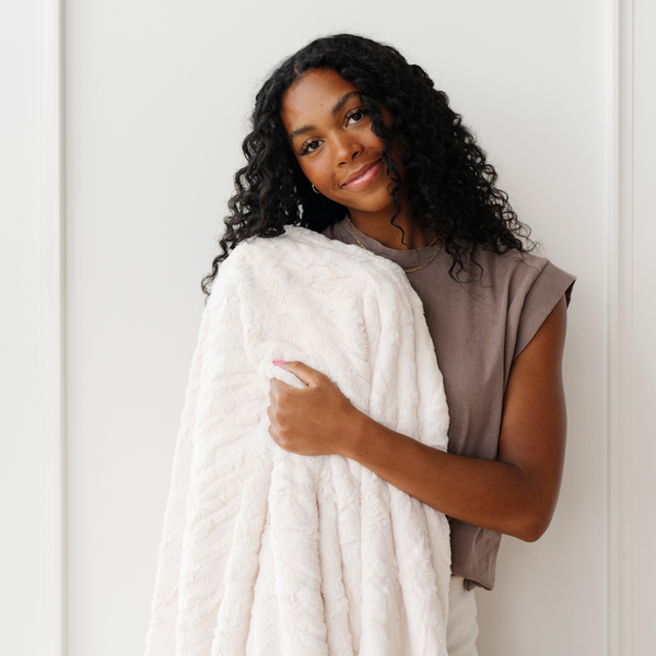 A woman smiles and holds an ivory luxurious patterned faux fur throw blanket that is a Saranoni blanket. This ultra-soft, plush throw blanket adds a touch of elegance and warmth to any space, perfect as a soft throw blanket or fluffy blanket for lounging. Ideal for those seeking a high-quality artificial fur blanket or luxury blanket, this versatile and stylish soft blanket enhances both comfort and decor. Shop the best blankets and throws for ultimate relaxation and cozy vibes.