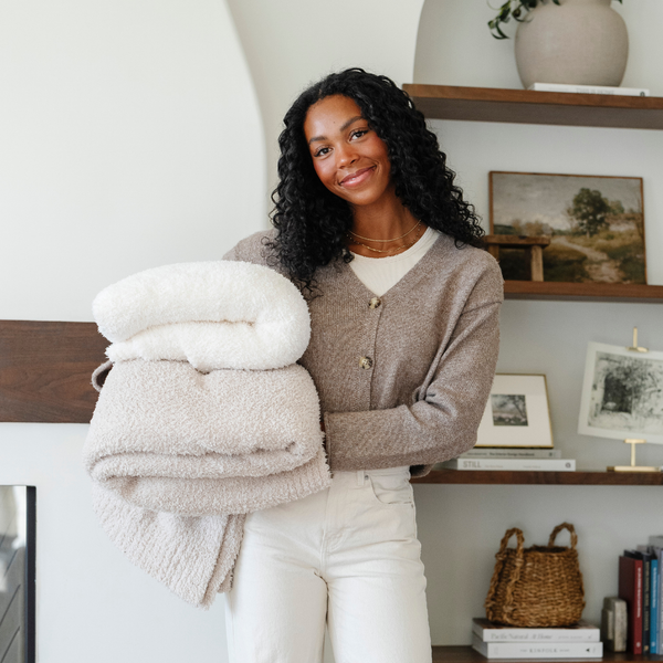 A woman holds a Bamboni® blanket by Saranoni. This ultra-soft blanket is a ivory blanket and made from a premium blend of bamboo rayon and poly microfiber, offering a lightweight, breathable, and fluffy feel. Perfect as a throw blanket, luxury blanket, plush throw, or soft blanket, it adds warmth and elegance to any living room, bedroom, or home décor setting. Designed for ultimate comfort, this buttery-soft Saranoni blanket is ideal for gifting. She also has a light tan Bamboni® blanket. 