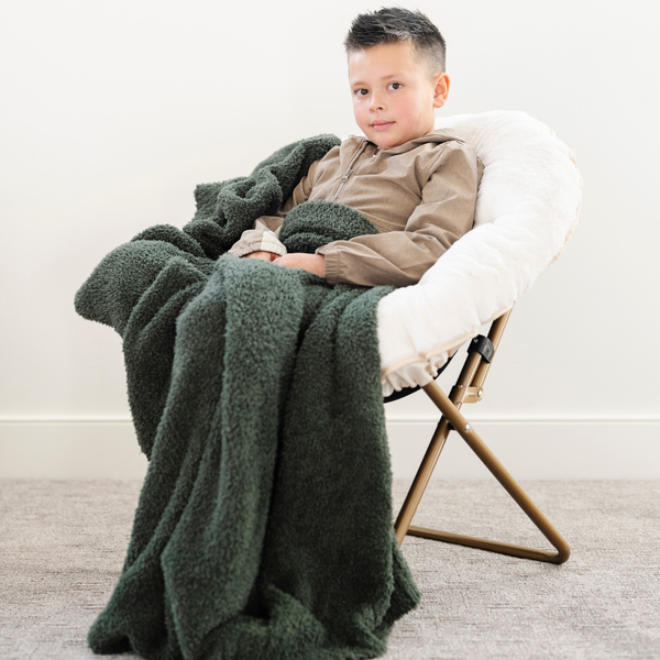 A little boy sits under a Bamboni® blanket by Saranoni. This ultra-soft blanket is a dark green blanket and made from a premium blend of bamboo rayon and poly microfiber, offering a lightweight, breathable, and fluffy feel. Perfect as a extra large blanket, luxury blanket, or soft blanket, it adds warmth and elegance to any living room, bedroom, or home décor setting. Designed for ultimate comfort, this buttery-soft Saranoni blanket is ideal for gifting. Enjoy this cozy blanket for years to come!