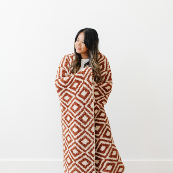 A woman holds a luxurious double-layer Bamboni® blanket by Saranoni, featuring a chic burnt orange and cream geometric pattern. This ultra-soft blanket is crafted from bamboo rayon and poly microfiber, making it lightweight, breathable, and fluffy. Ideal as a cozy throw blanket, luxury blanket, plush throw, soft blanket, or stylish home accent, this Saranoni blanket brings warmth, comfort, and timeless elegance to any living room or bedroom.