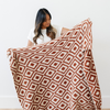 A woman holds up a luxurious double-layer Bamboni® blanket by Saranoni, featuring a chic burnt orange and cream geometric pattern. This ultra-soft blanket is crafted from bamboo rayon and poly microfiber, making it lightweight, breathable, and fluffy. Ideal as a cozy throw blanket, luxury blanket, plush throw, soft blanket, or stylish home accent, this Saranoni blanket brings warmth, comfort, and timeless elegance to any living room or bedroom.