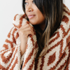 A woman holds a luxurious double-layer Bamboni® blanket by Saranoni, featuring a chic burnt orange and cream geometric pattern. This ultra-soft blanket is crafted from bamboo rayon and poly microfiber, making it lightweight, breathable, and fluffy. Ideal as a cozy blanket, extra large blanket, luxury blanket, soft blanket, or stylish home accent, this Saranoni blanket brings warmth, comfort, and timeless elegance to any living room or bedroom.