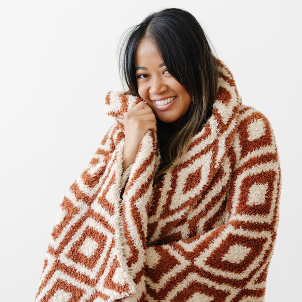 A smiling woman holds a luxurious double-layer Bamboni® blanket by Saranoni, featuring a chic burnt orange and cream geometric pattern. This ultra-soft blanket is crafted from bamboo rayon and poly microfiber, making it lightweight, breathable, and fluffy. Ideal as a cozy blanket, extra large blanket, luxury blanket, soft blanket, or stylish home accent, this Saranoni blanket brings warmth, comfort, and timeless elegance to any living room or bedroom.