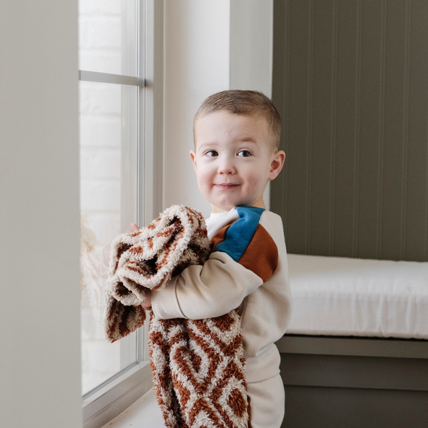 A little boy holds a double-layer Bamboni® baby blanket. The plush, breathable blanket featuring a chic burnt orange and cream geometric pattern, offering ultimate softness and warmth. Made from Saranoni’s signature stretchy, cloud-like Bamboni® fabric, this luxury baby blanket provides comfort for naps, tummy time, and on-the-go snuggles. The soft blanket is a mini blanket and Saranoni blanket. 