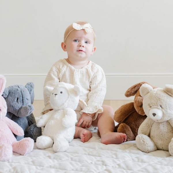 A delightful scene featuring a toddler sitting on a soft quilt surrounded by a collection of adorable stuffed animals, including a plush white lamb, a soft tan bear, a brown dog, and a gray elephant. The child, wearing a cute outfit and a bow headband, gazes curiously at the camera. Perfect for nurturing imaginative play and providing comfort, these cuddly toys create a warm and inviting atmosphere in a cozy nursery setting.