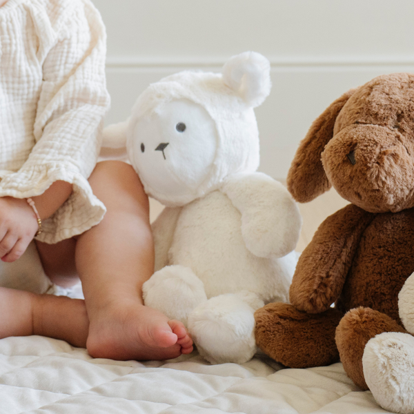 A charming close-up of a toddler sitting on a soft playmat, cuddling a plush white lamb stuffed animal with ears and a sweet expression. Next to the lamb is a soft brown stuffed dog. Perfect for nurturing imaginative play and cuddly moments in a cozy nursery setting.