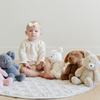 A delightful scene featuring a toddler sitting on a soft quilt surrounded by a collection of adorable stuffed animals, including a plush white lamb, a soft tan bear, a brown dog, and a gray elephant. The child, wearing a cute outfit and a bow headband, gazes curiously at the camera. Perfect for nurturing imaginative play and providing comfort, these cuddly toys create a warm and inviting atmosphere in a cozy nursery setting.