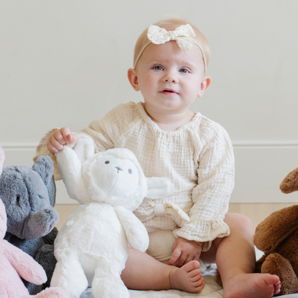 A joyful toddler sits on a soft quilt, proudly holding up a plush white lamb stuffed animal. Dressed in a cozy outfit and a cute bow headband, the child gazes at the camera with a cheerful expression. Surrounded by an array of adorable stuffed animals, including a gray elephant and a brown dog, this scene perfectly captures the warmth and comfort of childhood play, highlighting the importance of cuddly toys for nurturing imagination and joy.