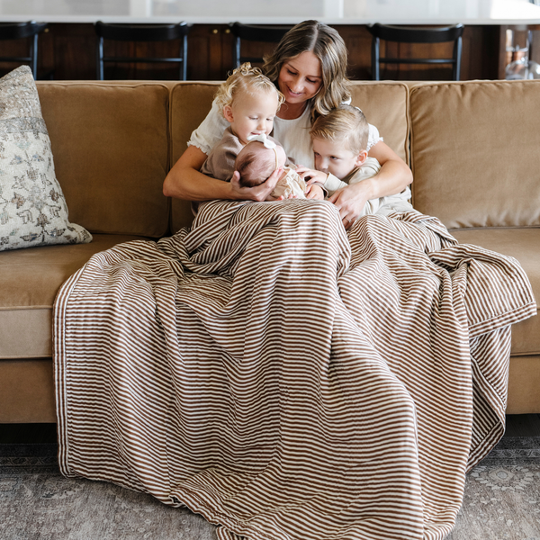 A mother and her three little kids sit with a Saranoni XL muslin quilt in a soft copper and white striped print. This luxury blanket features four layers of ultra-soft bamboo muslin, creating an airy yet cozy feel. Designed with a stitched quilted pattern and a one-inch border, this breathable blanket is perfect for muslin lovers looking for extra warmth and comfort. Made from bamboo fabric, this soft muslin quilt is a lightweight quilt that offers a cozy, neutral home décor accent. 