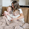 A mother and her kids sit with a Saranoni XL muslin quilt in a soft copper and white striped print. This luxury blanket features four layers of ultra-soft bamboo muslin, creating an airy yet cozy feel. Designed with a stitched quilted pattern and a one-inch border, this breathable blanket is perfect for muslin lovers looking for extra warmth and comfort. Made from bamboo fabric, this soft muslin quilt is a lightweight quilt that offers a cozy, neutral home décor accent. 