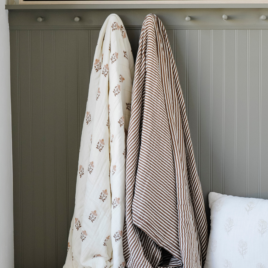 Two gorgeous Saranoni XL muslin quilts. One in a soft copper and white striped print and the other with a soft floral print. This luxury blanket features four layers of ultra-soft bamboo muslin, creating an airy yet cozy feel. Designed with a stitched quilted pattern and a one-inch border, this breathable blanket is perfect for muslin lovers looking for extra warmth and comfort. Made from bamboo fabric, this soft muslin quilt is a lightweight quilt that offers a cozy, neutral home décor accent. 