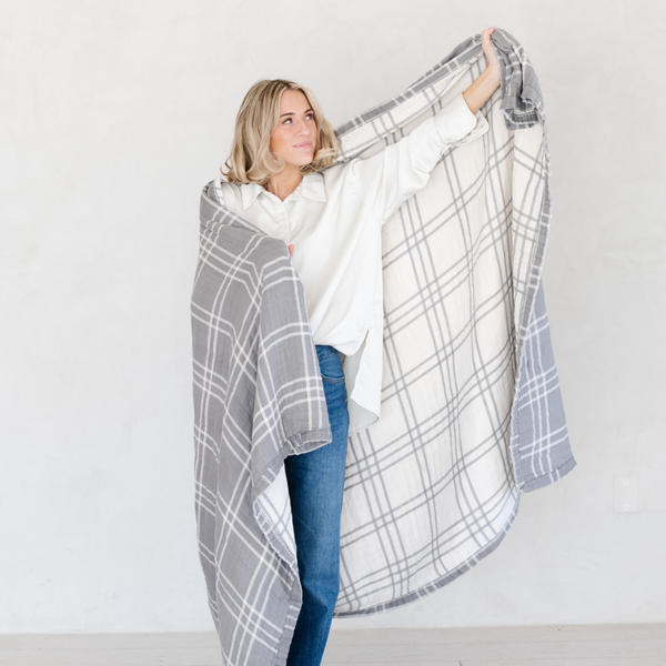 A woman wrapped in a gray and white Saranoni XL muslin quilt in a soft geometric print. This luxury blanket features four layers of ultra-soft bamboo muslin, creating an airy yet cozy feel. Designed with a stitched quilted pattern and a one-inch border, this breathable blanket is perfect for muslin lovers looking for extra warmth and comfort. Made from bamboo fabric, this soft muslin quilt is a lightweight quilt that offers a cozy, home décor accent. The softest blanket and a Saranoni blanket. 