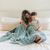 A mother sits with her son and daughter while holding a Luxury Extra Large Oversized Throw Eucalyptus Colored Lush Saranoni Blanket. The soft blanket is an extra large throw blanket and has a soft faux fur feeling.