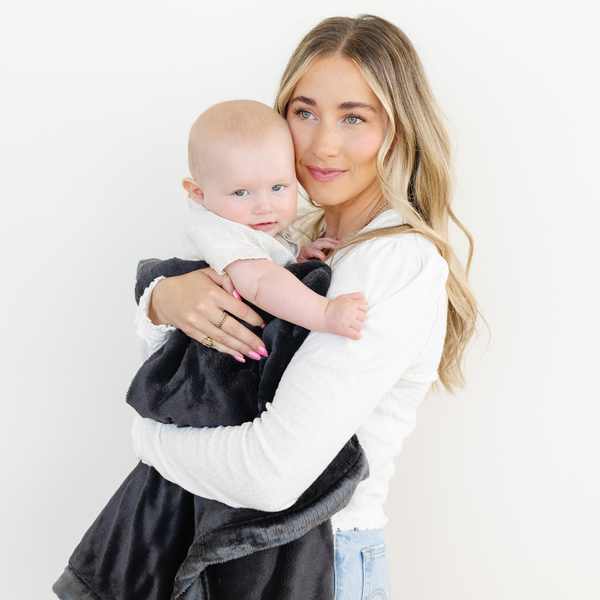 Mom stands holding baby boy snuggled with Charcoal lush blanket. 