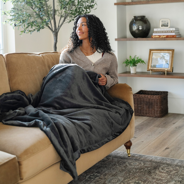 A woman sits on a couch with a Charcoal Lush XL Saranoni blanket, designed for oversized comfort and featuring a buttery soft, plush texture, on her lap. This luxurious Saranoni blanket is perfect for cozy evenings, reading, or home decor. The Saranoni blanket adds warmth, style, and unmatched softness, making it an ideal gift or essential for ultimate relaxation. The soft blanket is a luxury blanket and a Saranoni blanket. 