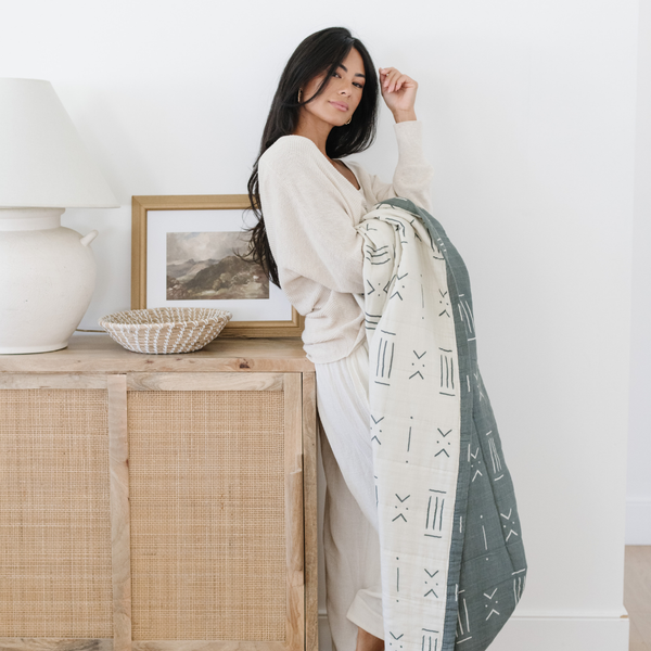 A woman holding a green and white Saranoni XL muslin quilt in a soft geometric print. This luxury blanket features four layers of ultra-soft bamboo muslin, creating an airy yet cozy feel. Designed with a stitched quilted pattern and a one-inch border, this breathable blanket is perfect for muslin lovers looking for extra warmth and comfort. Made from bamboo fabric, this soft muslin quilt is a lightweight quilt that offers a cozy, home décor accent. The softest blanket and a Saranoni blanket. 