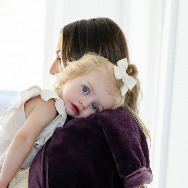 A mother holds her little girl who is laying on a Mini Aubergine (deep purple) Colored Lush Saranoni Blanket. The soft blanket is a small blanket and a baby blanket or toddler blanket.