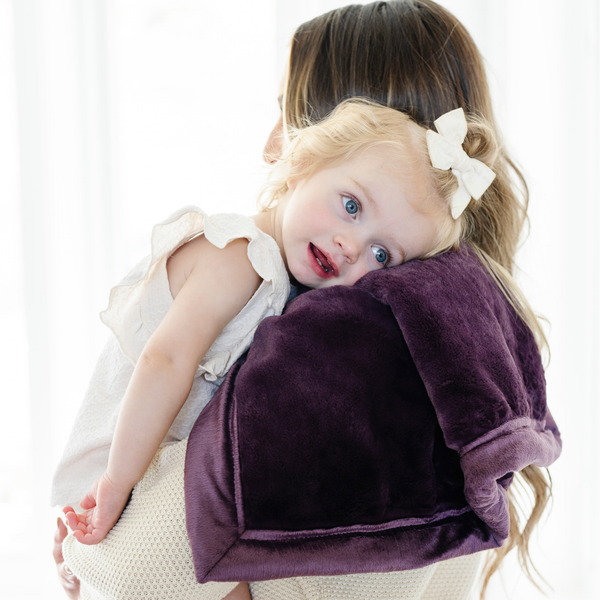 A mother holds her little girl who is laying on a Mini Aubergine (deep purple) Colored Lush Saranoni Blanket. The soft blanket is a small blanket and a baby blanket or toddler blanket.