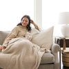 A woman smiles and sits with a luxurious ribbed Bamboni® blanket by Saranoni, the blanket is a very light tan. This ultra-soft blanket is crafted from bamboo rayon and poly microfiber, making it lightweight, breathable, and fluffy. Ideal as a cozy blanket, throw blanket, luxury blanket, soft blanket, or stylish home accent, this Saranoni blanket brings warmth, comfort, and timeless elegance to any living room or bedroom.