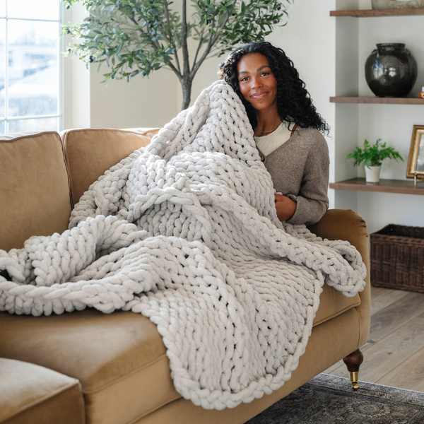 A woman smiles and sits on a couch with a soft, light gray colored Saranoni chunky knit throw blanket. The large knit Saranoni blanket, is made from 100% poly microfiber and drapes beautifully. This luxury throw blanket is both trendy and durable, making it the perfect addition to any space. Ideal for home decor enthusiasts and those seeking a cuddly, soft blanket, this chunky knit blanket combines style and comfort. Keywords: chunky knit throw, soft blanket, knit blanket, luxury throw blanket.