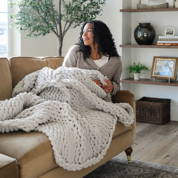 A woman sits on a couch with a soft, light gray colored Saranoni chunky knit throw blanket. The large knit Saranoni blanket, is made from 100% poly microfiber and drapes beautifully. This luxury throw blanket is both trendy and durable, making it the perfect addition to any space. Ideal for home decor enthusiasts and those seeking a cuddly, soft blanket, this chunky knit blanket combines style and comfort. Keywords: chunky knit throw, soft blanket, knit blanket, large knit blanket, luxury throw blanket.