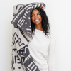 A smiling woman holds a luxurious double-layer Bamboni® blanket by Saranoni, featuring a chic black and cream geometric pattern. This ultra-soft blanket is crafted from bamboo rayon and poly microfiber, making it lightweight, breathable, and fluffy. Ideal as a cozy throw blanket, luxury blanket, plush throw, soft blanket, or stylish home accent, this Saranoni blanket brings warmth, comfort, and timeless elegance to any living room or bedroom.