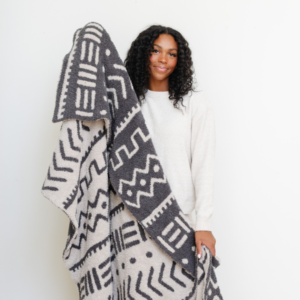 A woman smiles and holds a luxurious double-layer Bamboni® blanket by Saranoni, featuring a chic black and cream geometric pattern. This ultra-soft blanket is crafted from bamboo rayon and poly microfiber, making it lightweight, breathable, and fluffy. Ideal as a cozy throw blanket, luxury blanket, plush throw, soft blanket, or stylish home accent, this Saranoni blanket brings warmth, comfort, and timeless elegance to any living room or bedroom.