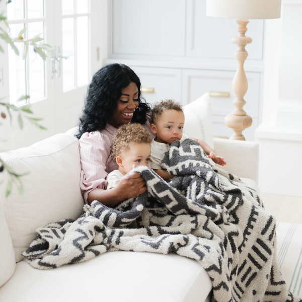 A mother and her two little boys sit with a luxurious double-layer Bamboni® blanket by Saranoni, featuring a chic black and cream geometric pattern. This ultra-soft blanket is crafted from bamboo rayon and poly microfiber, making it lightweight, breathable, and fluffy. Ideal as a cozy throw blanket, luxury blanket, plush throw, soft blanket, or stylish home accent, this Saranoni blanket brings warmth, comfort, and timeless elegance to any living room or bedroom.