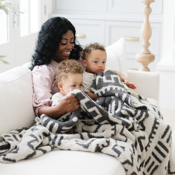 A mother sits with her with her two little boys and a luxurious double-layer Bamboni® blanket by Saranoni, featuring a chic black and cream geometric pattern. This ultra-soft blanket is crafted from bamboo rayon and poly microfiber, making it lightweight, breathable, and fluffy. Ideal as a cozy blanket, extra large blanket, luxury blanket, soft blanket, or stylish home accent, this Saranoni blanket brings warmth, comfort, and timeless elegance to any living room or bedroom.