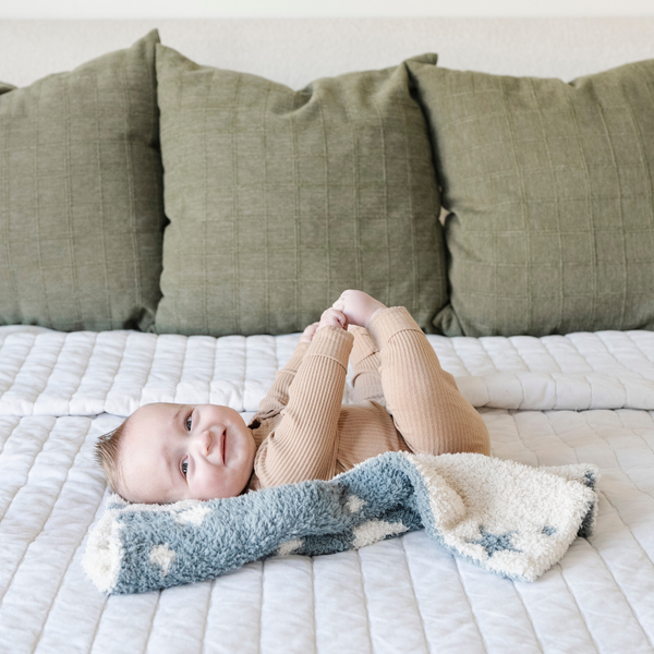 A smiling baby boy with a double-layer Bamboni® baby blanket. The plush, breathable blanket features a two-tone blue and cream star design, offering ultimate softness and warmth. Made from Saranoni’s signature stretchy, cloud-like Bamboni® fabric, this luxury baby blanket provides comfort for naps, tummy time, and on-the-go snuggles. The perfect blend of warmth and breathability, it’s a must-have for parents seeking high-quality, ultra-soft blankets. The soft blanket is a mini blanket and Saranoni blanket. 