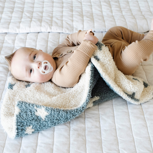 A baby boy lays with a double-layer Bamboni® baby blanket. The plush, breathable blanket features a two-tone blue and cream star design, offering ultimate softness and warmth. Made from Saranoni’s signature stretchy, cloud-like Bamboni® fabric, this luxury baby blanket provides comfort for naps, tummy time, and on-the-go snuggles. The perfect blend of warmth and breathability, it’s a must-have for parents seeking high-quality, ultra-soft blankets. The soft blanket is a mini blanket and Saranoni blanket. 