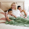 A mother and her kids smile and sit with a Bamboni® blanket by Saranoni. This ultra-soft blanket is a light green blanket and made from a premium blend of bamboo rayon and poly microfiber, offering a lightweight, breathable, and fluffy feel. Perfect as a extra large blanket, luxury blanket, or soft blanket, it adds warmth and elegance to any living room, bedroom, or home décor setting. Designed for ultimate comfort, this buttery-soft Saranoni blanket is ideal for gifting. 