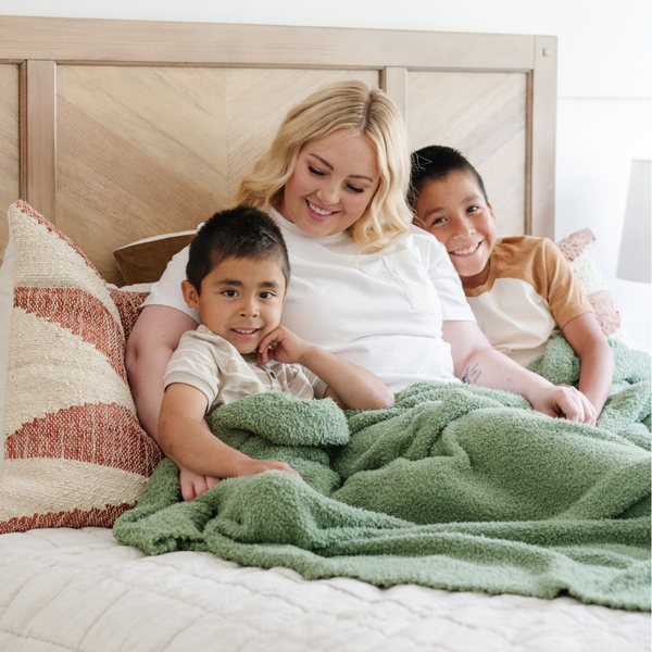 A mother and her kids sit under a Bamboni® blanket by Saranoni. This ultra-soft blanket is a light green blanket and made from a premium blend of bamboo rayon and poly microfiber, offering a lightweight, breathable, and fluffy feel. Perfect as a extra large blanket, luxury blanket, or soft blanket, it adds warmth and elegance to any living room, bedroom, or home décor setting. Designed for ultimate comfort, this buttery-soft Saranoni blanket is ideal for gifting. Enjoy this cozy blanket for years to come!