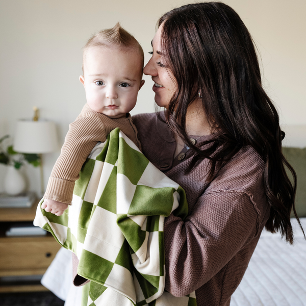 A mother smiles holds her baby boy and a olive and cream checkered minky stretch blanket. The soft blanket is a luxury blanket and a Saranoni blanket. This cozy blanket is also a receiving blanket and is a cute and cozy baby blanket. This stretchy blanket is a great nursery item. 