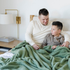 A father and son sit with a Bamboni® blanket by Saranoni. This ultra-soft blanket is a light green blanket and made from a premium blend of bamboo rayon and poly microfiber, offering a lightweight, breathable, and fluffy feel. Perfect as a extra large blanket, luxury blanket, or soft blanket, it adds warmth and elegance to any living room, bedroom, or home décor setting. Designed for ultimate comfort, this buttery-soft Saranoni blanket is ideal for gifting. Enjoy this cozy blanket for years to come!