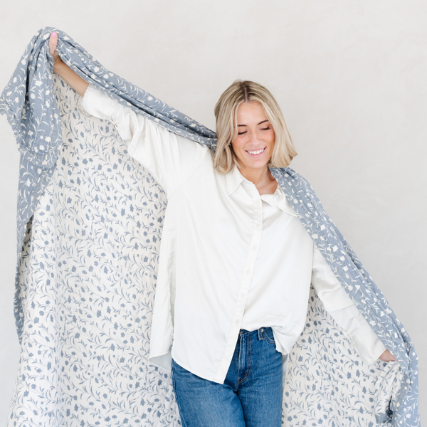 A woman holding a blue and white Saranoni XL muslin quilt in a soft floral print. This luxury blanket features four layers of ultra-soft bamboo muslin, creating an airy yet cozy feel. Designed with a stitched quilted pattern and a one-inch border, this breathable blanket is perfect for muslin lovers looking for extra warmth and comfort. Made from bamboo fabric, this soft muslin quilt is a lightweight quilt that offers a cozy, home décor accent. The softest blanket and a Saranoni blanket. 