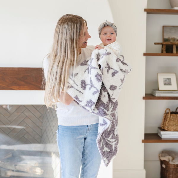 A mother smiles and holds her baby girl and a double-layer Bamboni® baby blanket. The plush, breathable blanket features a two-tone purple and white floral design, offering ultimate softness and warmth. Made from Saranoni’s signature stretchy, cloud-like Bamboni® fabric, this luxury baby blanket provides comfort for naps, tummy time, and on-the-go snuggles. The soft blanket is a receiving blanket and Saranoni blanket. 