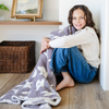 A little girl smiles and holds a double-layer Bamboni® blanket. The plush, breathable blanket features a two-tone purple and white floral design, offering ultimate softness and warmth. Made from Saranoni’s signature stretchy, cloud-like Bamboni® fabric, this luxury kids blanket provides comfort for naps and on-the-go snuggles. The soft blanket is a toddler blanket and Saranoni blanket. 