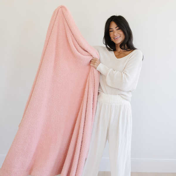 A woman smiles and holds up a Bamboni® blanket by Saranoni. This ultra-soft blanket is a peach blanket and made from a premium blend of bamboo rayon and poly microfiber, offering a lightweight, breathable, and fluffy feel. Perfect as a extra large blanket, luxury blanket, or soft blanket, it adds warmth and elegance to any living room, bedroom, or home décor setting. Designed for ultimate comfort, this buttery-soft Saranoni blanket is ideal for gifting. Enjoy this cozy blanket for years to come!