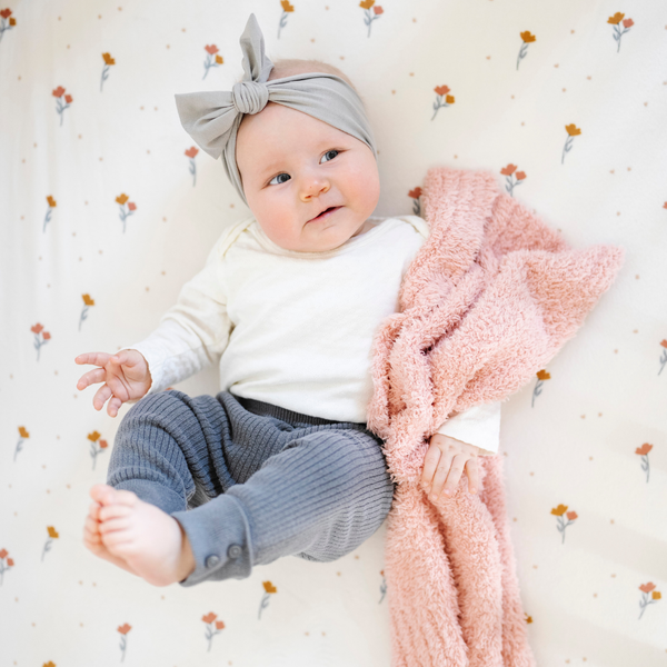 A baby girl is laying down with a peach blanket that is a Bamboni® blanket, offering ultimate softness and warmth. Made from Saranoni’s signature stretchy, cloud-like Bamboni® fabric, this luxury baby blanket provides comfort for naps, tummy time, and on-the-go snuggles. The soft blanket is a mini blanket and Saranoni blanket. 