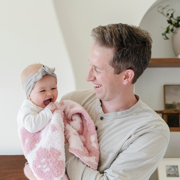 A father smiles and holds his baby girl who is holding a double-layer Bamboni® baby blanket. The plush, breathable blanket features a two-tone light pink and pink floral design, offering ultimate softness and warmth. Made from Saranoni’s signature stretchy, cloud-like Bamboni® fabric, this luxury baby blanket provides comfort for naps, tummy time, and on-the-go snuggles. The soft blanket is a mini blanket and Saranoni blanket. 