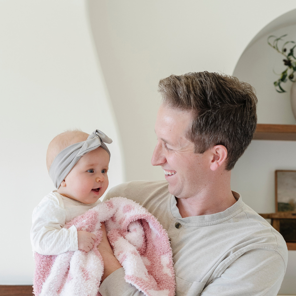 A father holds his baby girl who is holding a double-layer Bamboni® baby blanket. The plush, breathable blanket features a two-tone light pink and pink floral design, offering ultimate softness and warmth. Made from Saranoni’s signature stretchy, cloud-like Bamboni® fabric, this luxury baby blanket provides comfort for naps, tummy time, and on-the-go snuggles. The soft blanket is a mini blanket and Saranoni blanket. 