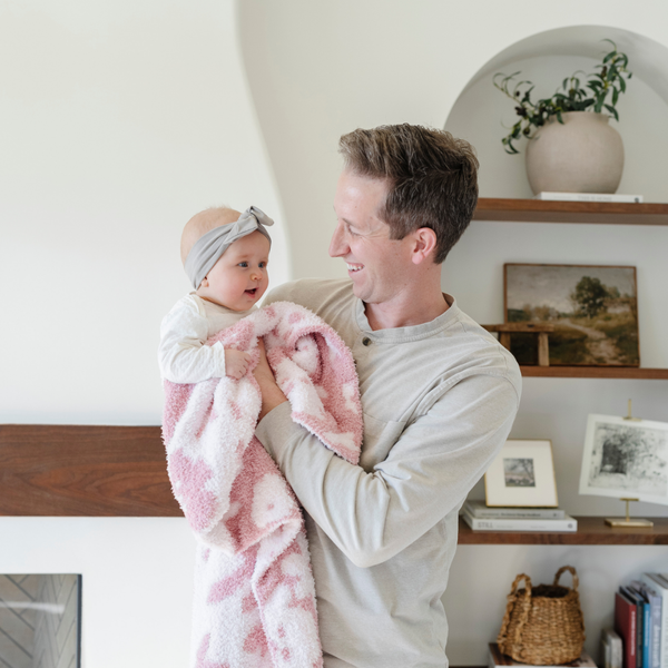 A father smiles and holds his baby girl who is holding a double-layer Bamboni® baby blanket. The plush, breathable blanket features a two-tone light pink and pink floral design, offering ultimate softness and warmth. Made from Saranoni’s signature stretchy, cloud-like Bamboni® fabric, this luxury baby blanket provides comfort for naps, tummy time, and on-the-go snuggles. The soft blanket is a receiving blanket and Saranoni blanket. 