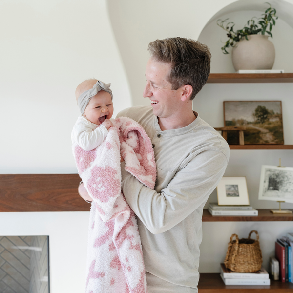 A father smiles and holds his happy baby girl who is holding a double-layer Bamboni® baby blanket. The plush, breathable blanket features a two-tone light pink and pink floral design, offering ultimate softness and warmth. Made from Saranoni’s signature stretchy, cloud-like Bamboni® fabric, this luxury baby blanket provides comfort for naps, tummy time, and on-the-go snuggles. The soft blanket is a receiving blanket and Saranoni blanket. 